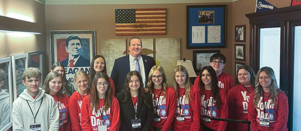 A group of teenage students wearing red, long-sleeved Youth Day shirts surround a state senator in his office