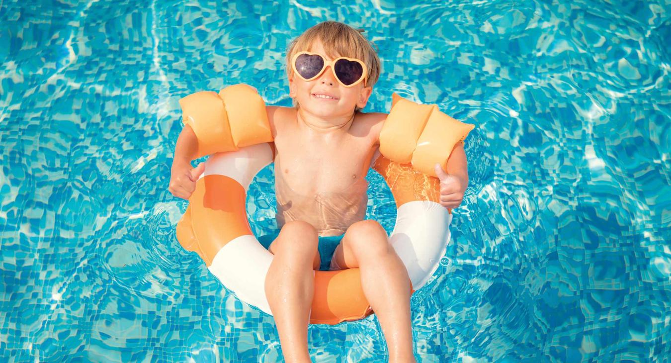 A young boy in an orange and white intertube floats in a swimming pool. He's wearing orange water wings, orange heart-shaped sunglasses and is looking up at the camera with a smile and two thumbs up.