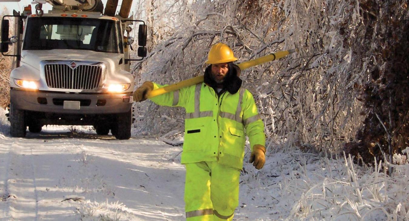 A winter scene with a lineworker in layers of warm safety gear walks on a snow-covered road. Behind him is a utility bucket truck carrying utility poles.