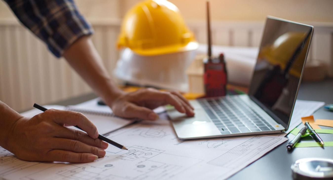 Contractor makring up plans while working on laptop. Hard hats and a walkie talkie are in the background