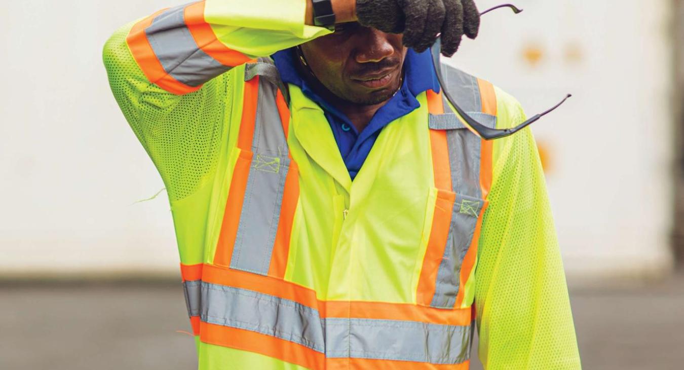 A worker in yellow hard hat and yellow reflective jacket wipes sweat from his brow